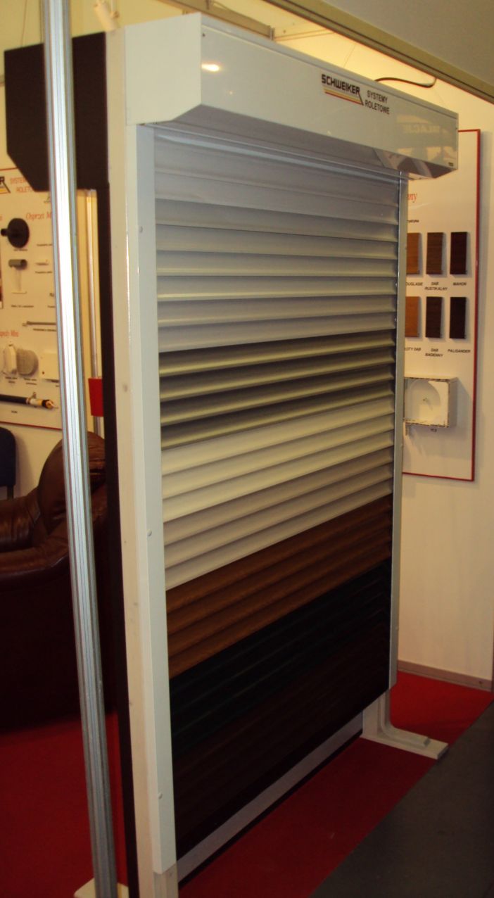 Wystawowa brama rolowana z lamelami w odcieniach szarości, brązu i czerni, prezentowana na tle ściany z próbkami kolorów i elementami sterowania.